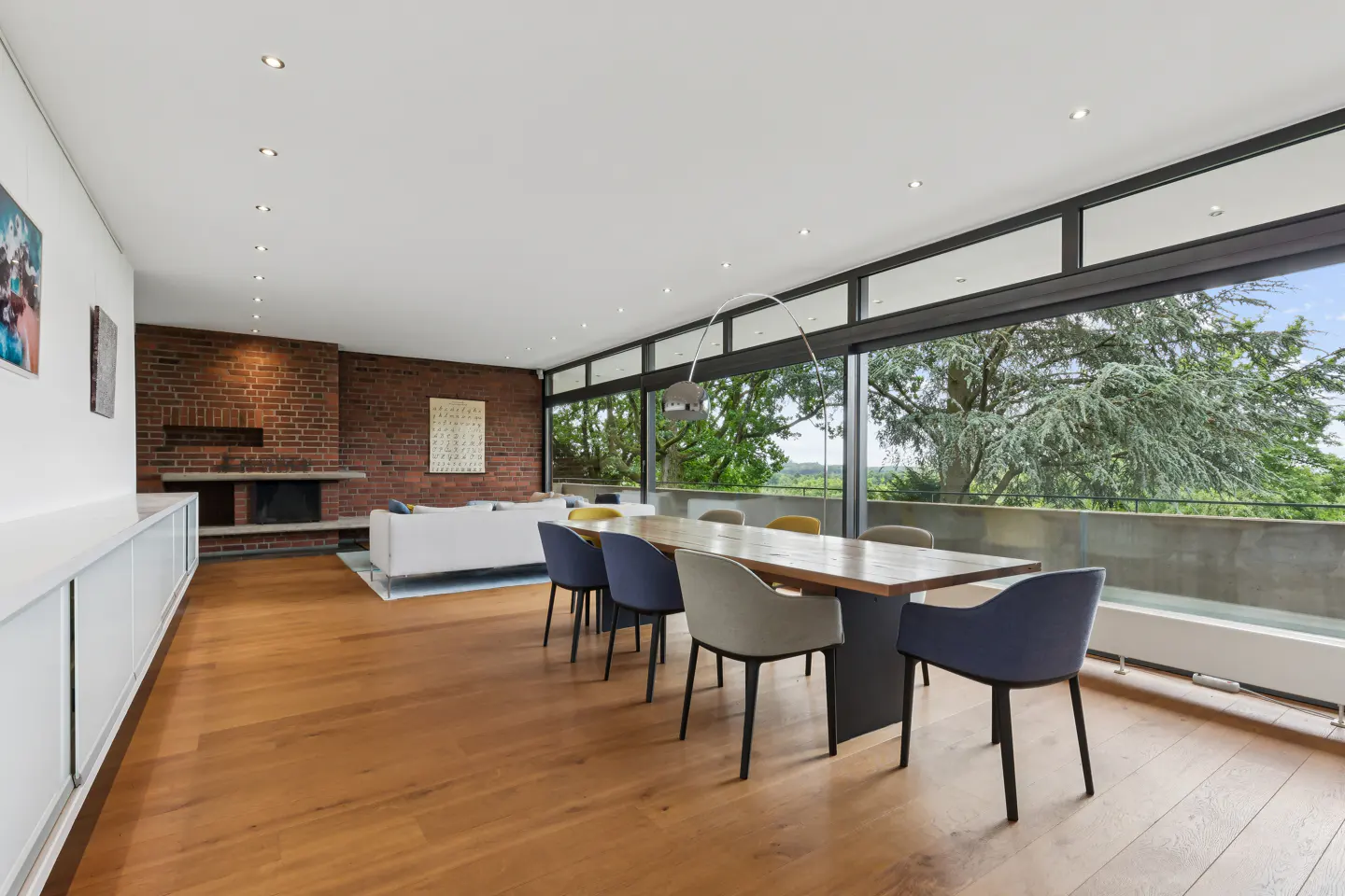 Modern dining area with wooden floors, a long table, colorful chairs, and a brick wall fireplace overlooking lush trees.