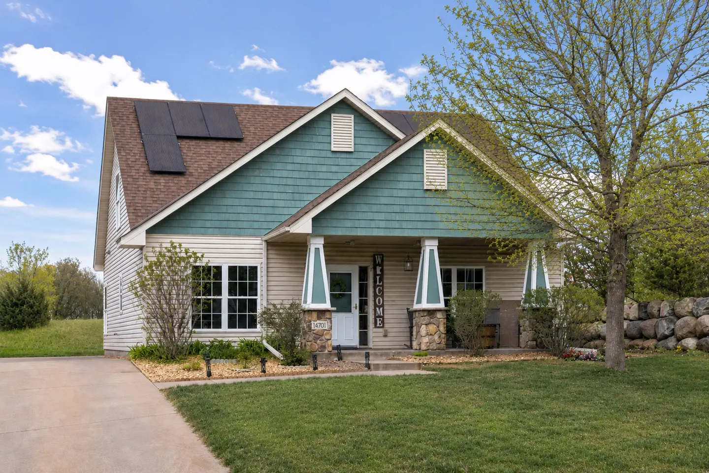 Tan house with green trim, solar panels, and a "Welcome" sign on the porch. Lawn and trees surround the house.