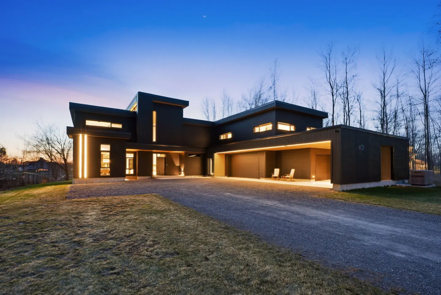 Modern black house with gravel driveway and warm lighting at dusk. Trees in the background.