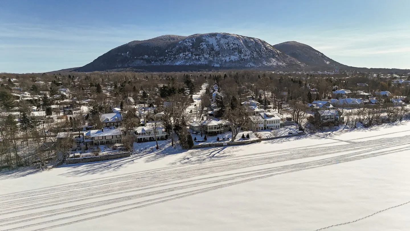 Aerial view of snow-covered homes near a frozen lake, with a large mountain in the background under a blue sky.