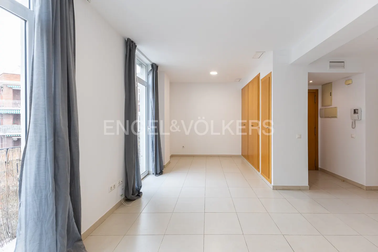 Bright, empty room with white walls, tile floor, and wooden closets. Gray curtains frame windows with a balcony view. Engel & Völkers logo visible.