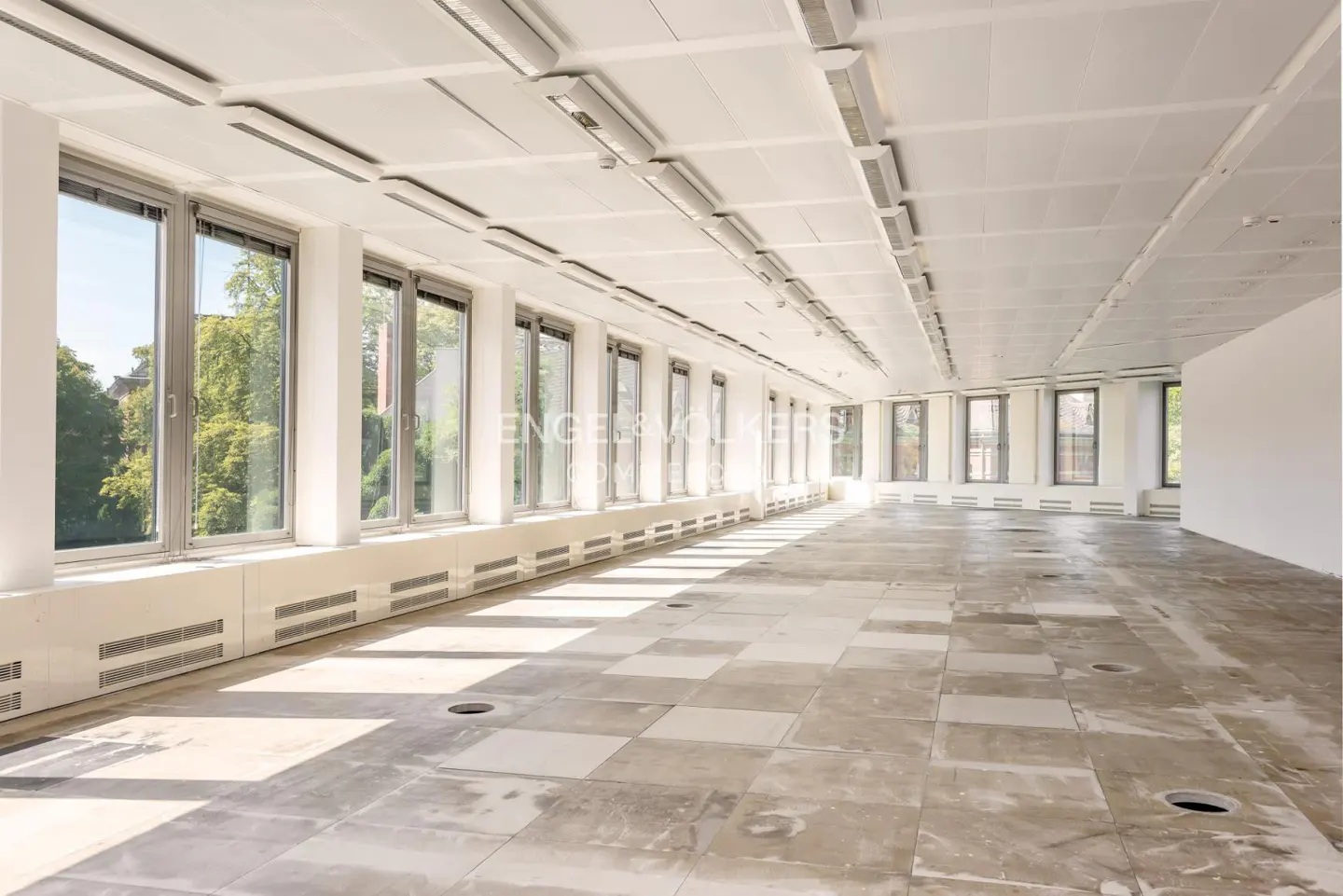 Bright, empty office space with large windows, white walls, and a tiled floor. Overhead lighting illuminates the room.