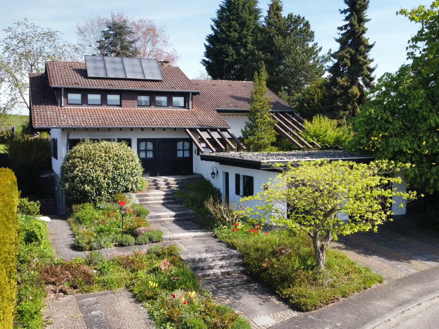 Two-story house with a red tile roof and solar panels. A stone walkway leads to the front door, surrounded by lush greenery and colorful flowers.