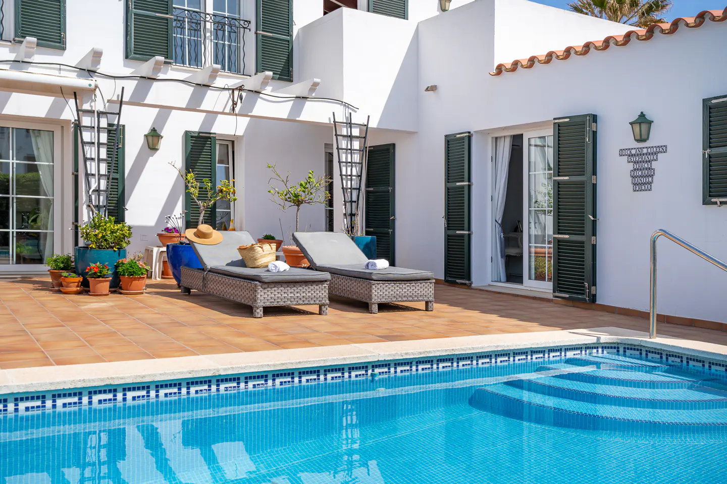 Outdoor patio with two lounge chairs, a pool with blue tiles, and a white building with green shutters.