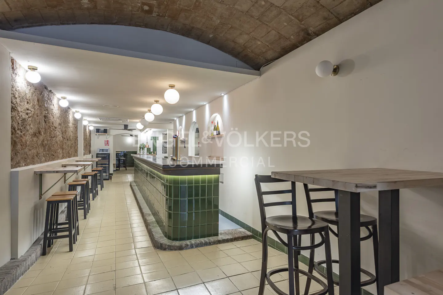 Interior of a commercial space with a green tiled bar, tables, stools, and exposed brick wall.