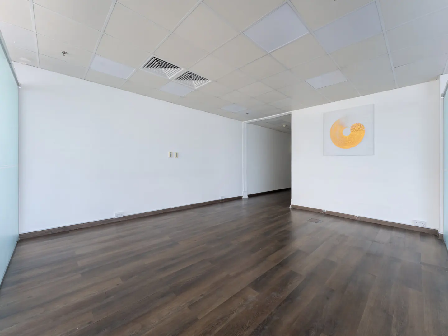 Empty office space with dark wood floors, white walls, and a white ceiling with square lights. A yellow circle art piece hangs on the wall.