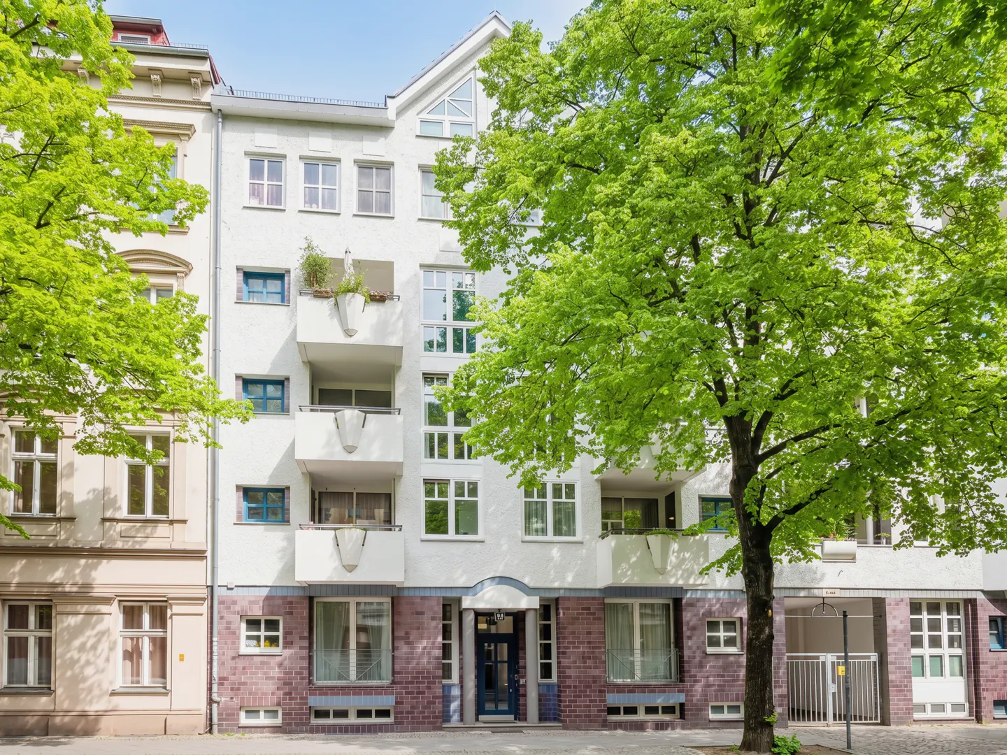 Exterior of a white apartment building with balconies and a brick facade, framed by green trees.