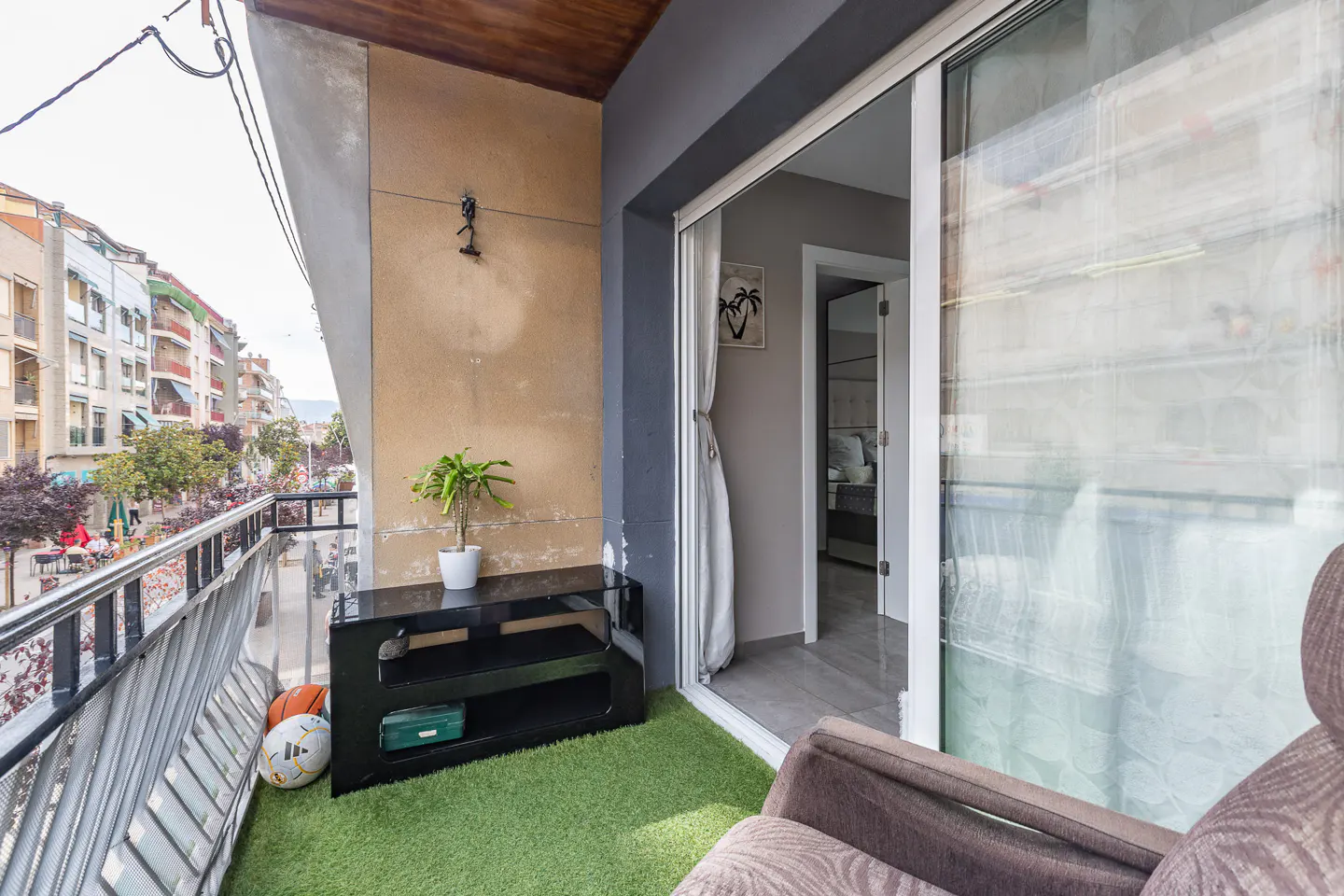 A balcony with artificial grass, a black console table with a plant, two balls, and a view of a city street.