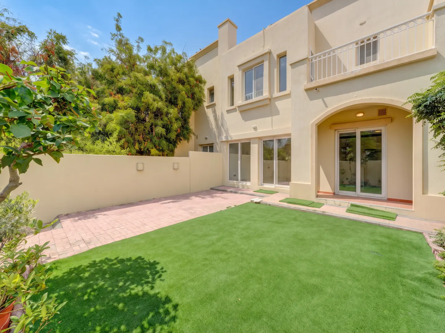 Backyard view of a two-story beige house with a green lawn, brick patio, and trees under a blue sky.