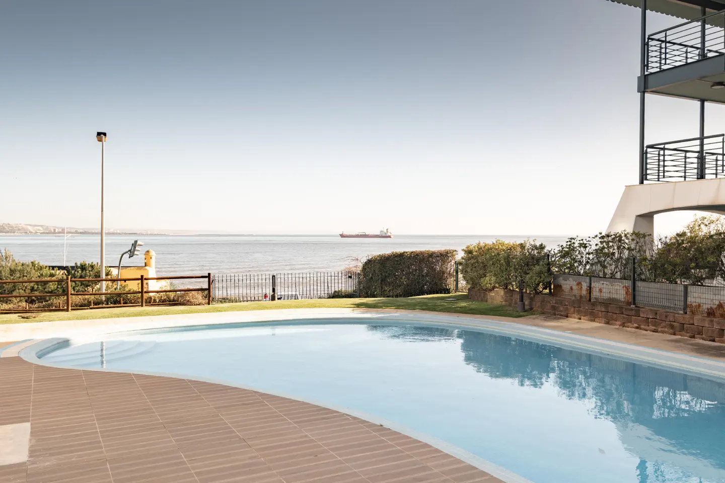 A light blue swimming pool with brown tile surround overlooks the ocean with a ship in the distance.