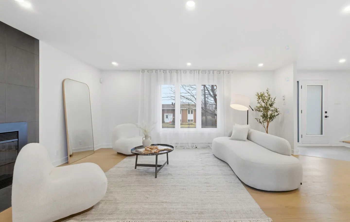 Bright living room with white walls, wood floors, and a gray rug. White furniture includes a curved sofa, chairs, and a black coffee table. A tall mirror leans against the wall.