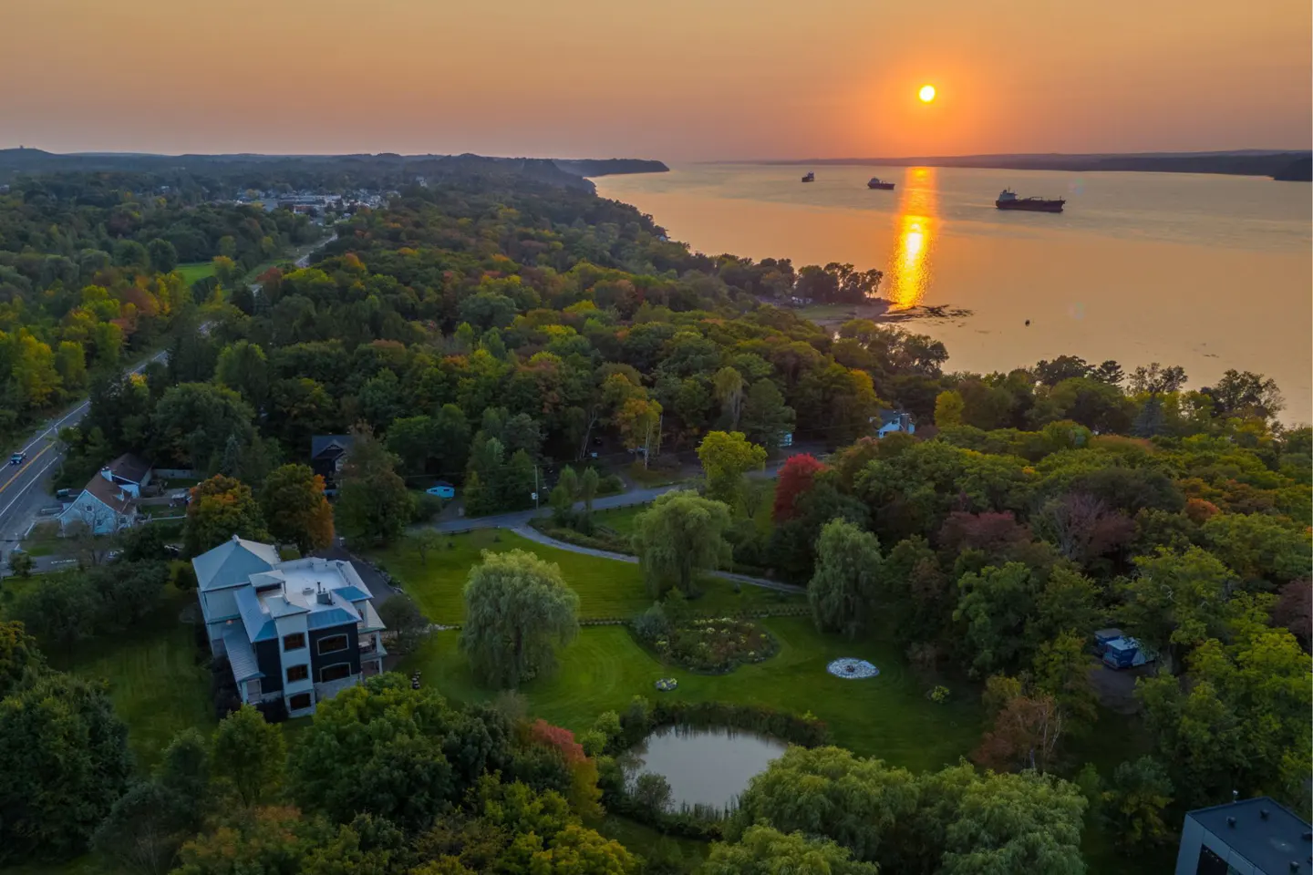 Aerial view of a modern house surrounded by trees, a pond, and a river at sunset.