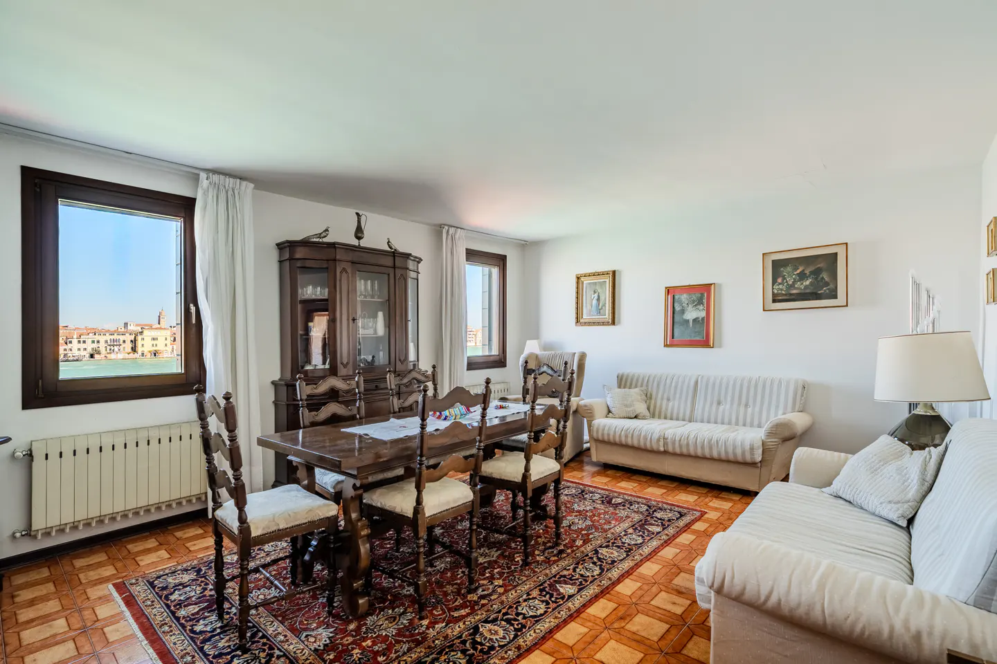 Bright living room with a wood dining table, chairs, and a china cabinet. Two sofas sit on a red patterned rug. A window shows a Venice canal view.
