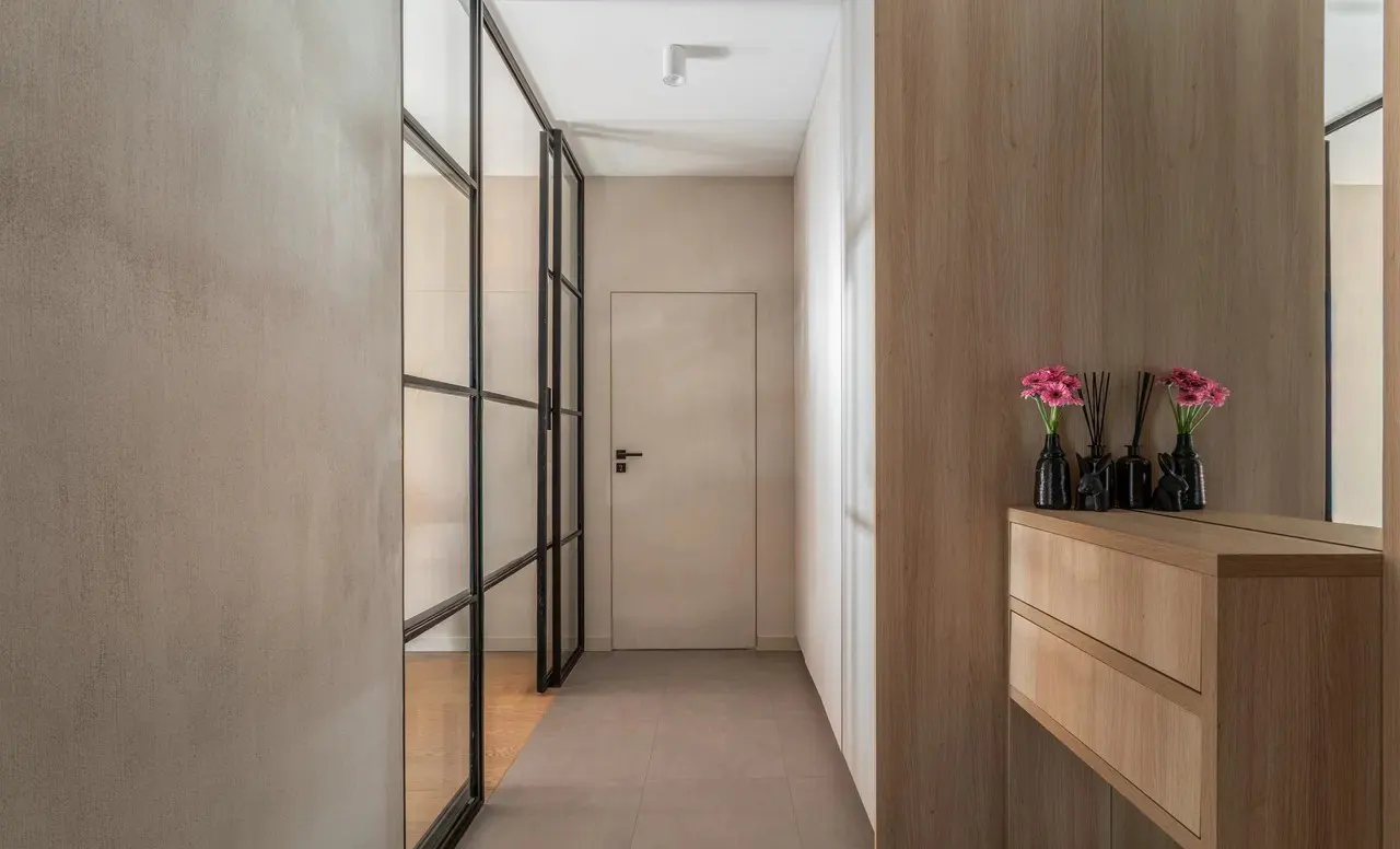 Hallway with a black-framed glass partition, light wood cabinets, and pink flowers in black vases.