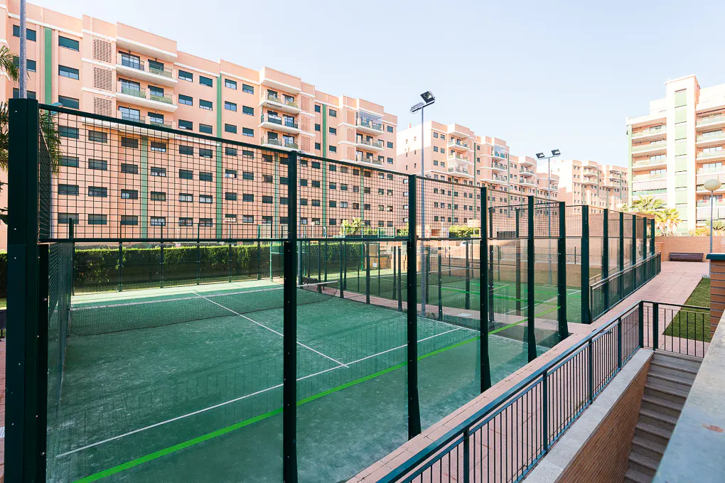 Outdoor paddle tennis courts with green surfaces and dark green fencing, set against a backdrop of peach-colored apartment buildings.
