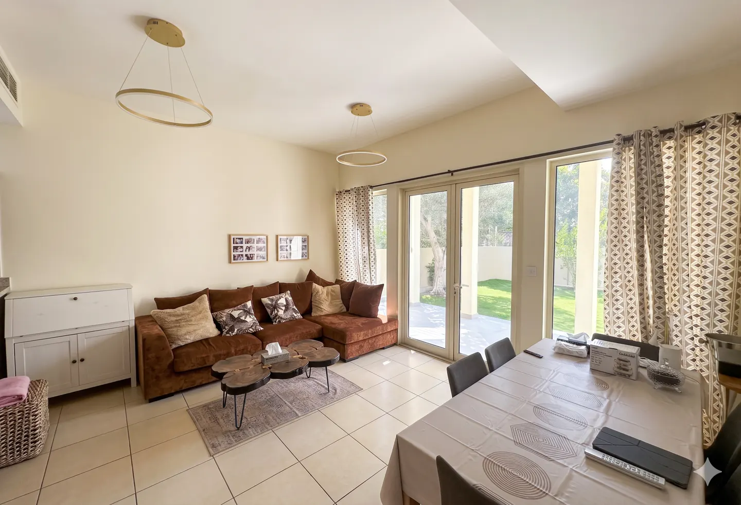 Living room with a brown sectional sofa, wood-slice coffee table, dining area, and glass doors leading to a garden.