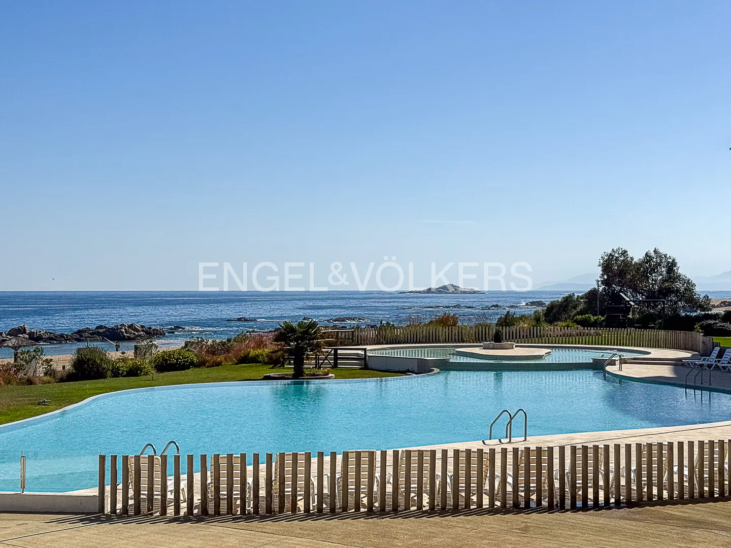 A large, blue swimming pool overlooks the ocean on a sunny day. A wooden fence surrounds the pool.