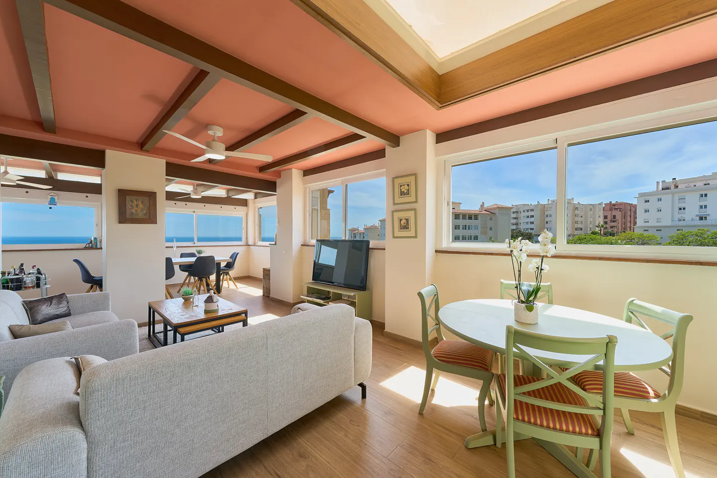 Bright living room with sofas, dining table, and large windows overlooking city and ocean. Pink ceiling with wooden beams and a ceiling fan.