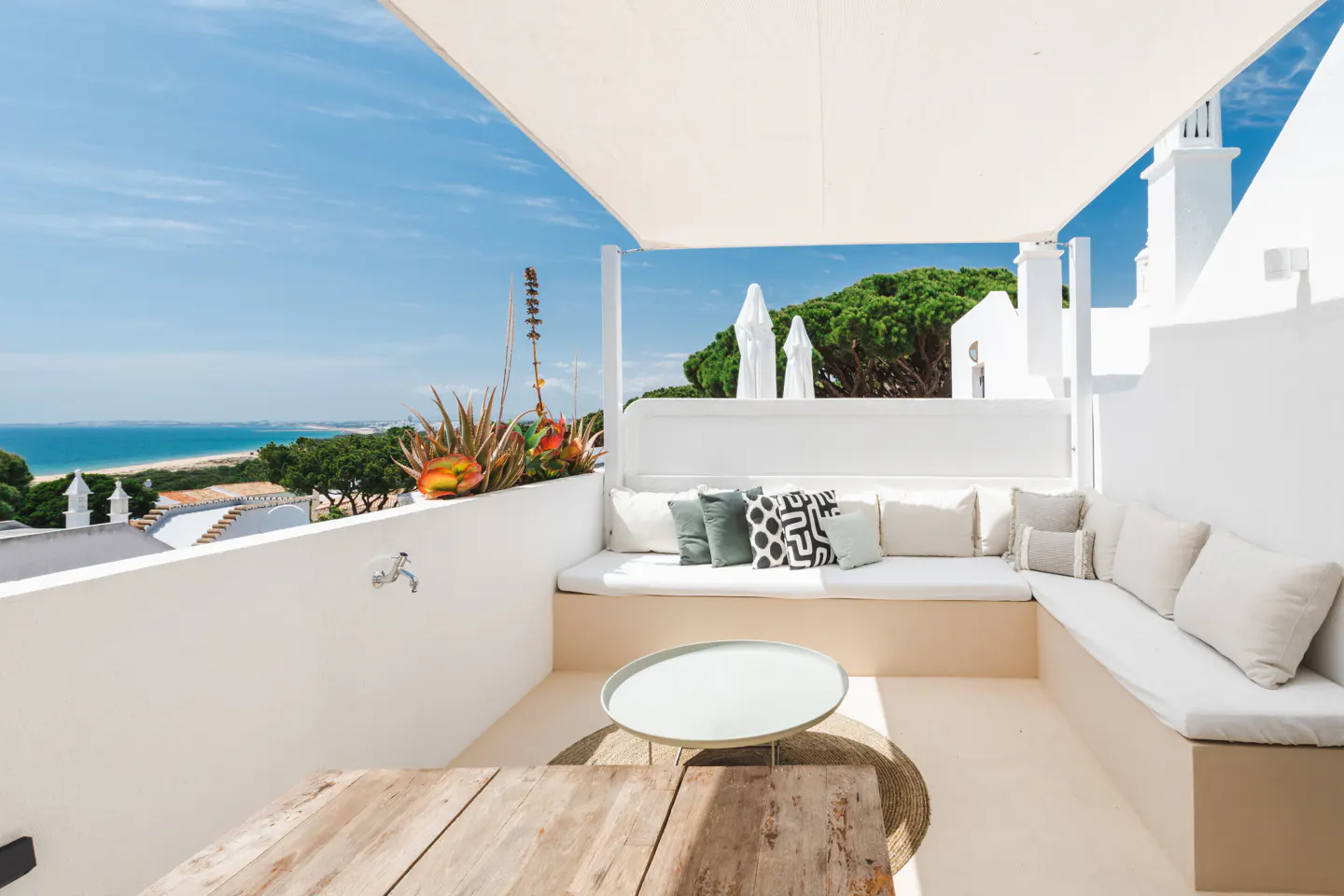 A bright, white rooftop patio with a built-in sofa, pillows, and a wooden table overlooking the ocean.
