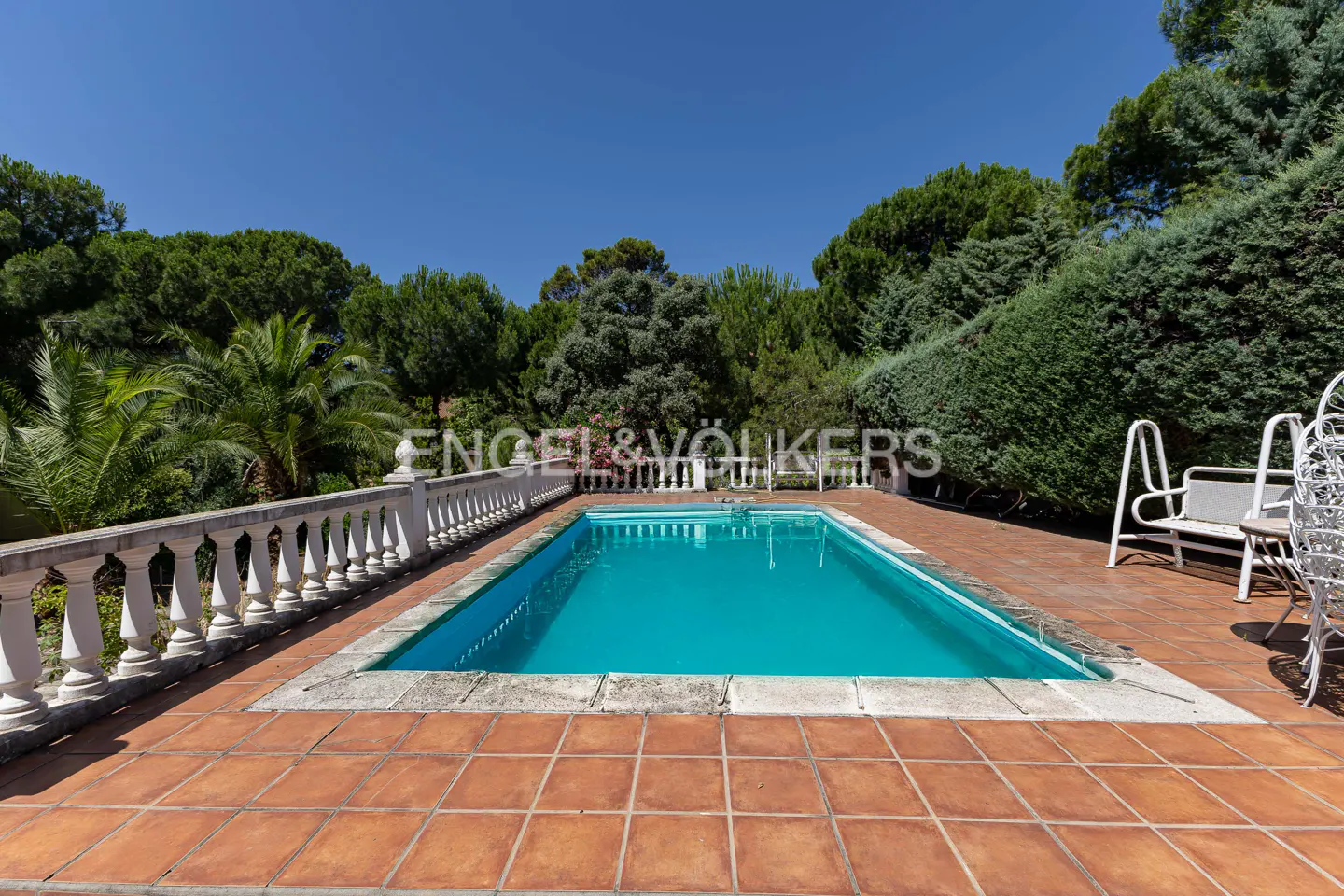 Outdoor pool with turquoise water, surrounded by terracotta tiles and a white balustrade. Green trees and a clear blue sky in the background.