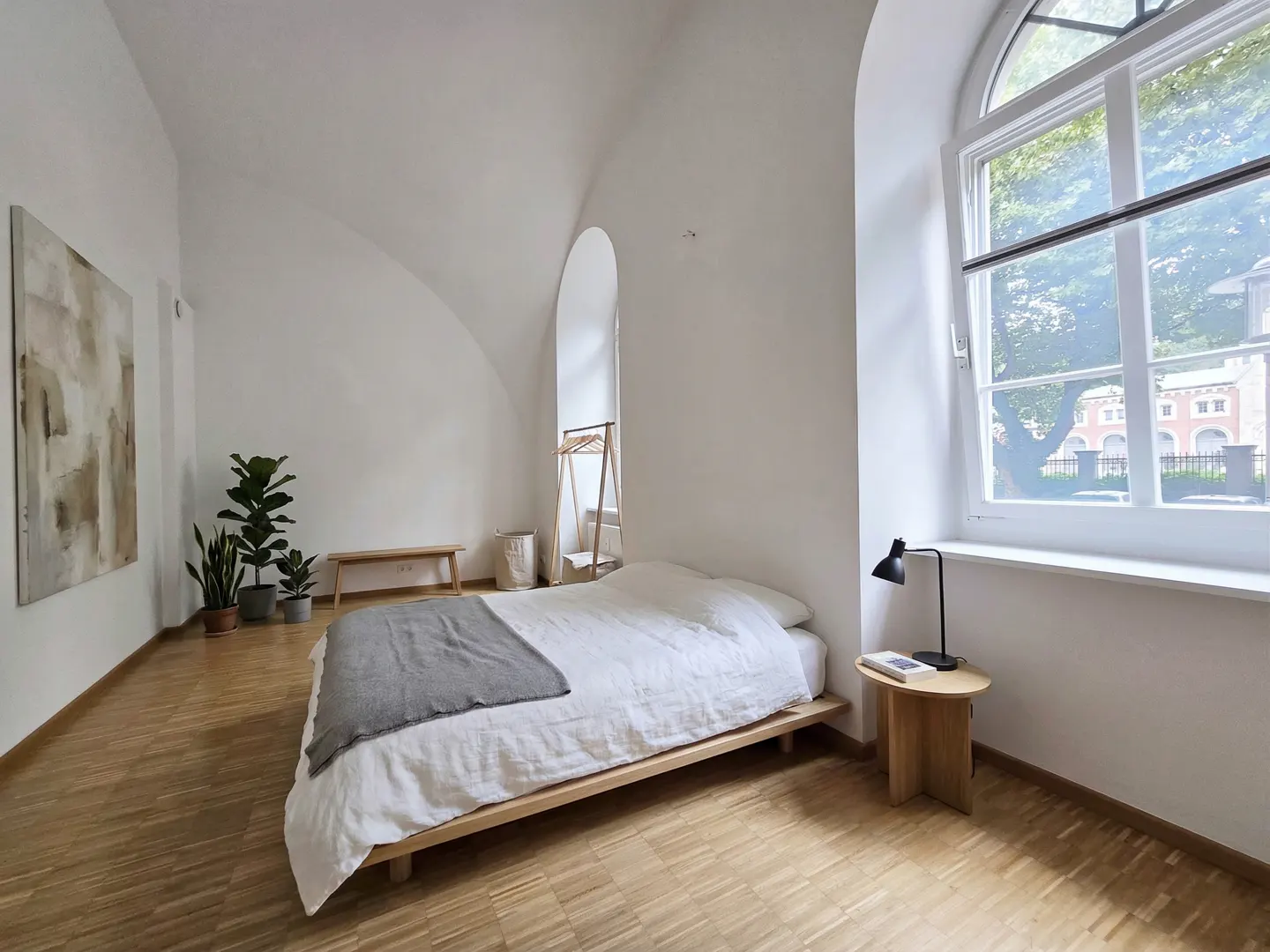 Bright bedroom with white walls, wood floors, and a large arched window. A bed with white linens and a gray blanket sits in the center. A black lamp is on a small table.