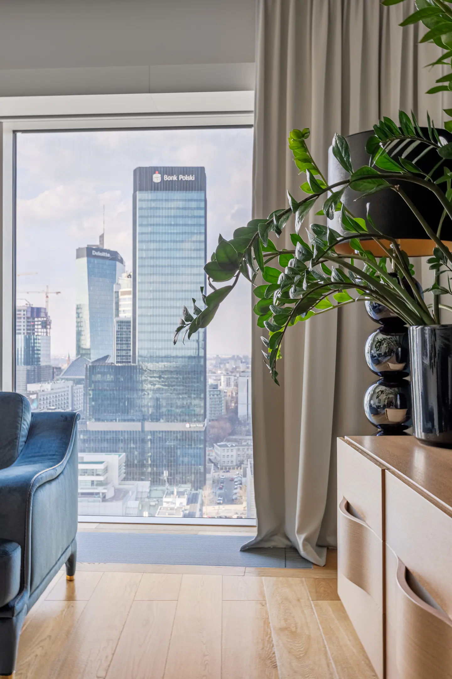 Apartment interior with a city view. A blue sofa sits near a large window overlooking skyscrapers. A plant and light wood cabinet are on the right.