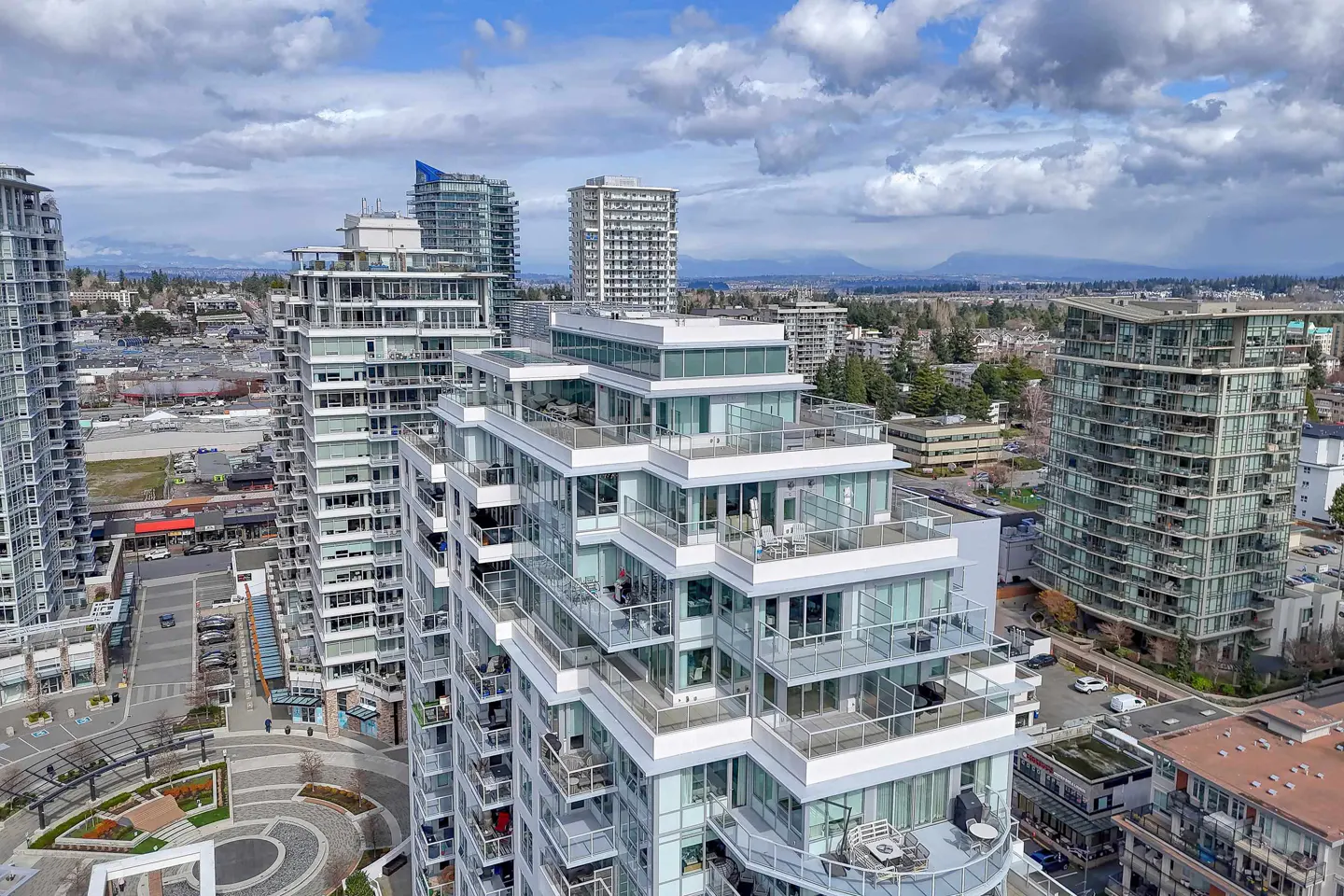 Aerial view of modern white high-rise condos with glass balconies, set against a backdrop of mountains and a cloudy blue sky.