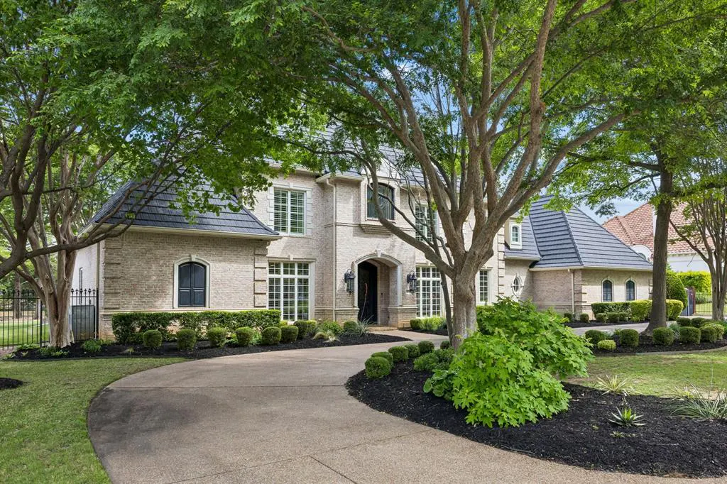 Beige brick home with a dark roof, surrounded by green trees and landscaping. A curved driveway leads to the front entrance.