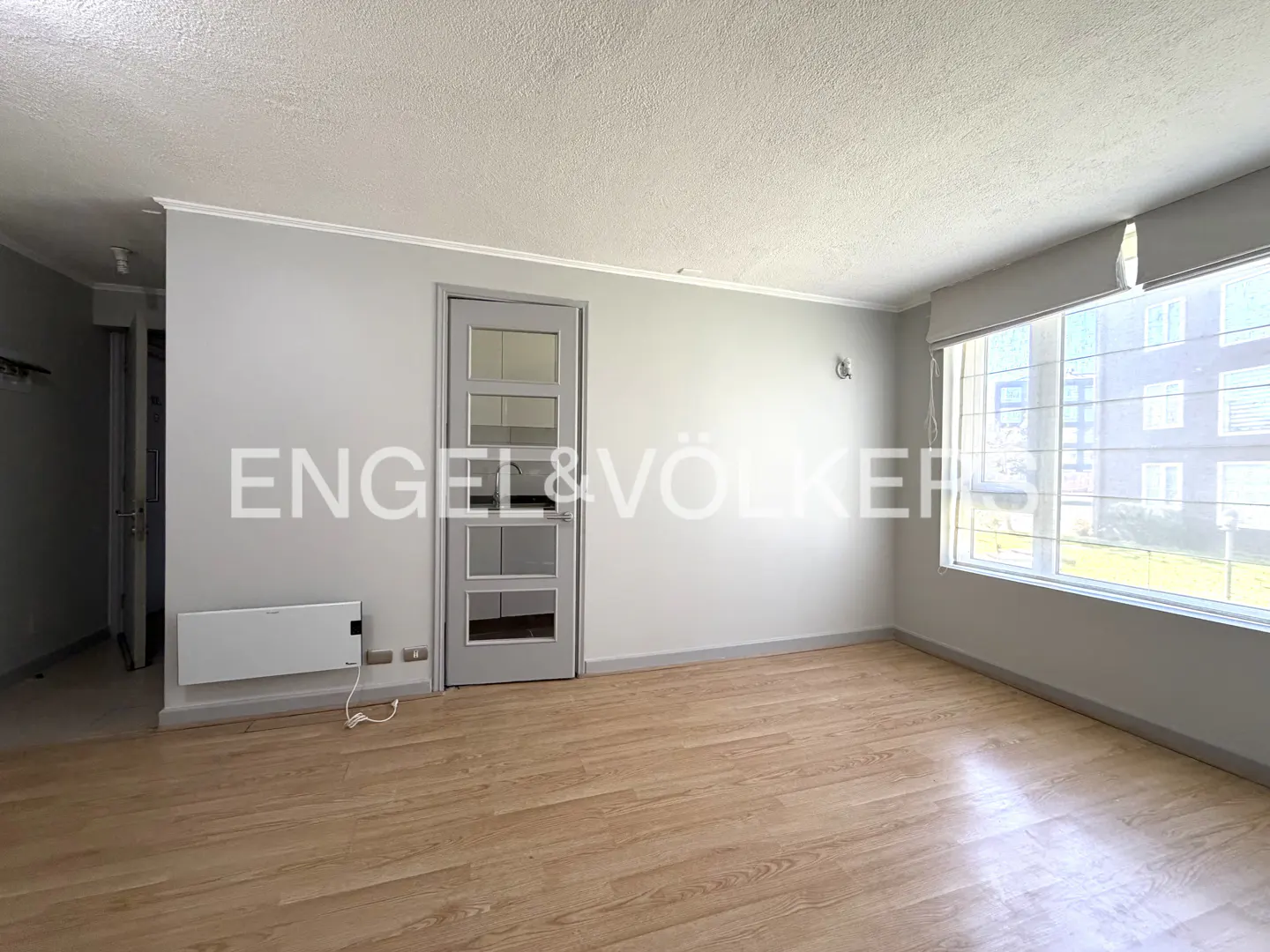 Empty room with light wood floors, gray walls, and a window with a view of an apartment building. Engel & Völkers logo on the wall.