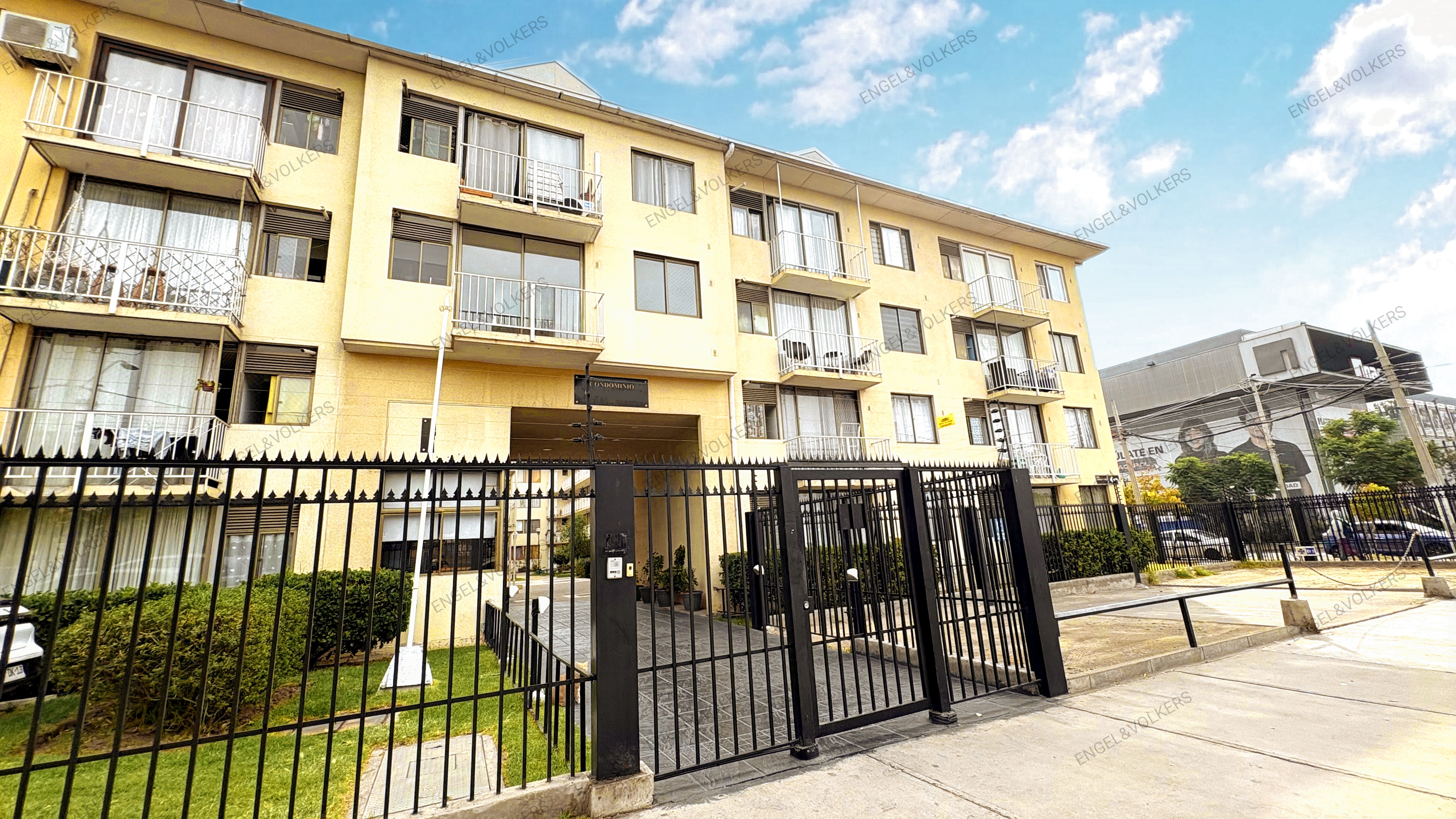 Exterior view of a yellow apartment building with black iron fence and gate. Balconies and windows are visible.
