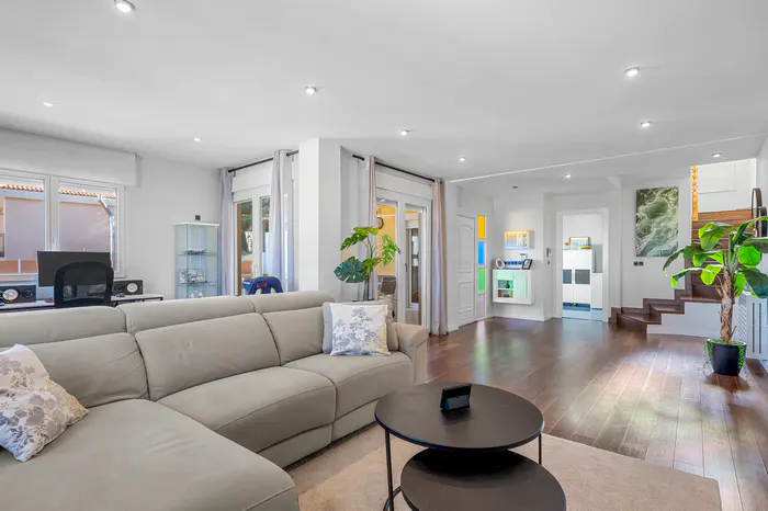 Bright living room with a beige sectional sofa, dark wood floors, and white walls. A staircase and potted plant are visible in the background.