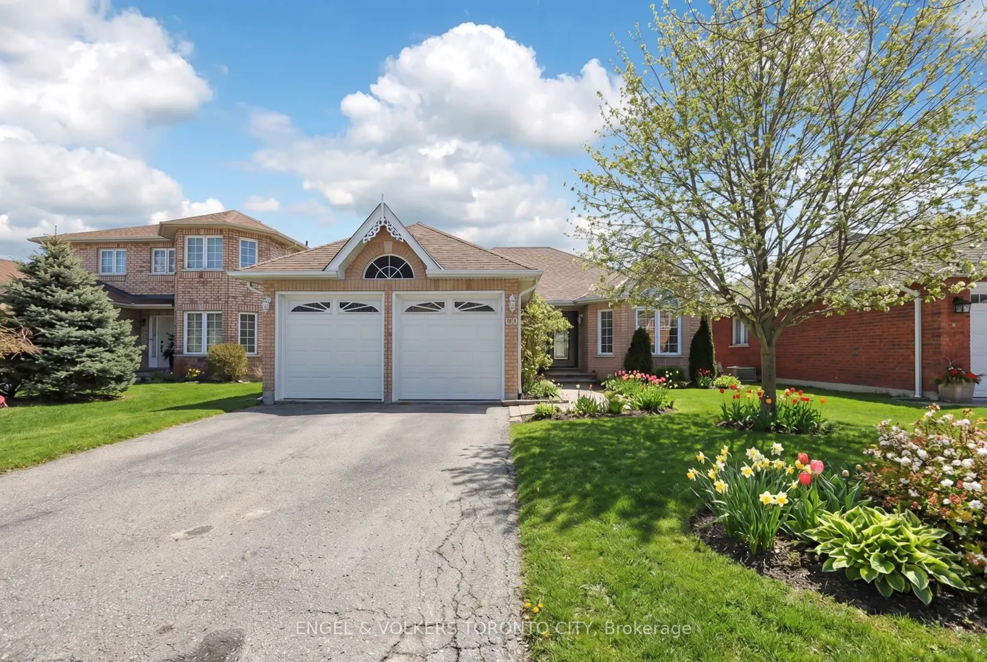 Exterior of a one-story brick house with a two-car garage, green lawn, and colorful flower garden on a sunny day.