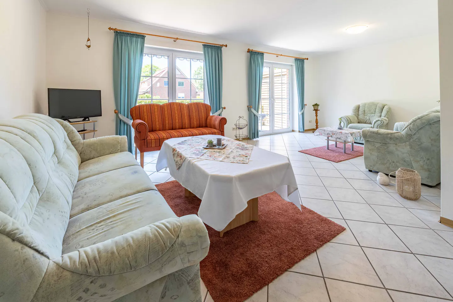 Bright living room with tile floors, sofas, armchairs, and a table with a white tablecloth. Blue curtains frame the windows.