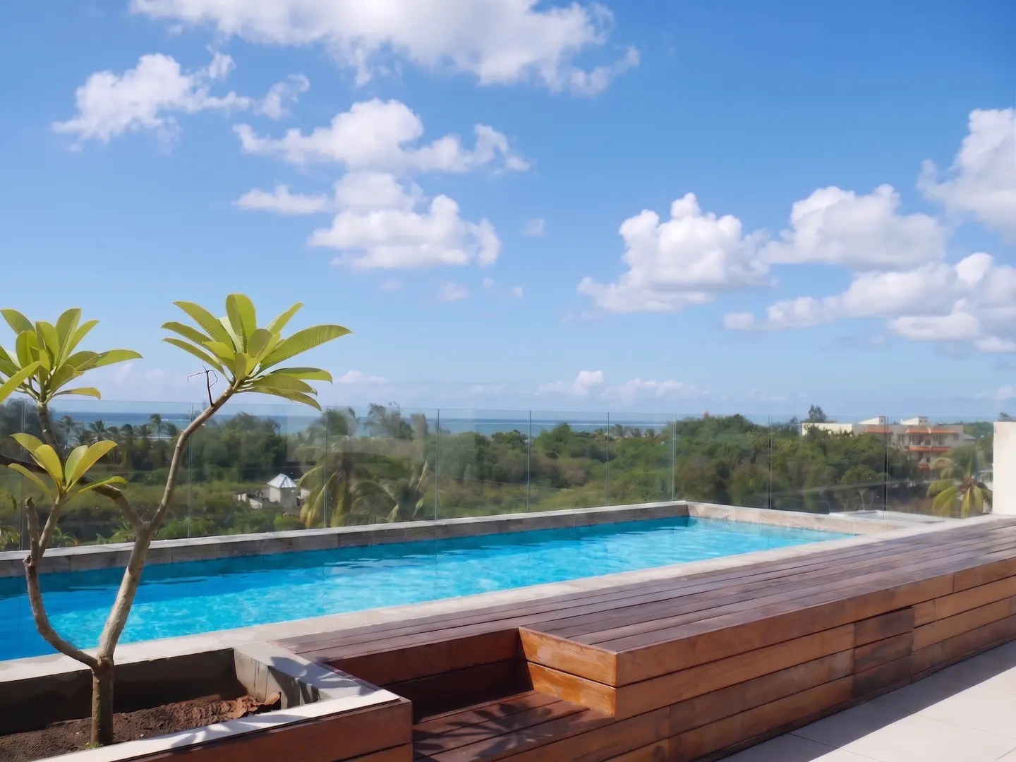 Rooftop pool with wooden deck and ocean view. Blue sky with white clouds above green trees. A small tree in a pot is in the foreground.