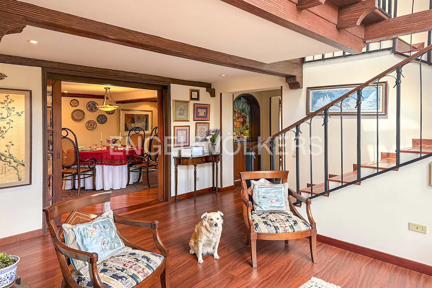 A bright foyer with wood floors, beams, and staircase. Two chairs face a dog sitting in the center. A dining room is visible through an open doorway.