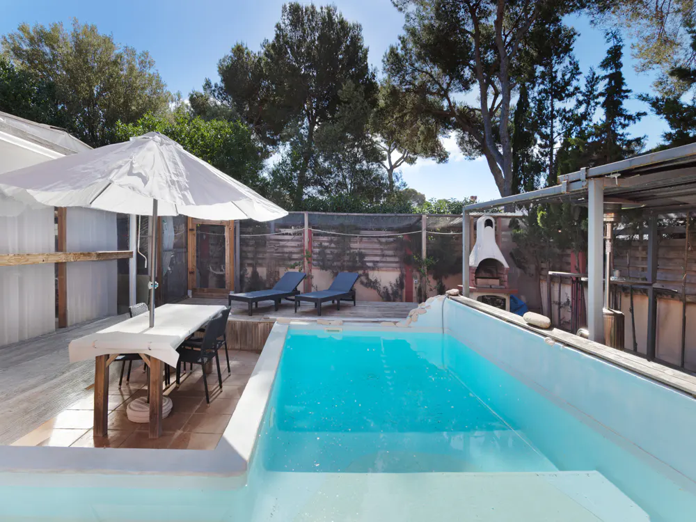 Outdoor pool area with turquoise water, lounge chairs, and a dining table under a white umbrella. Trees and a blue sky are visible in the background.