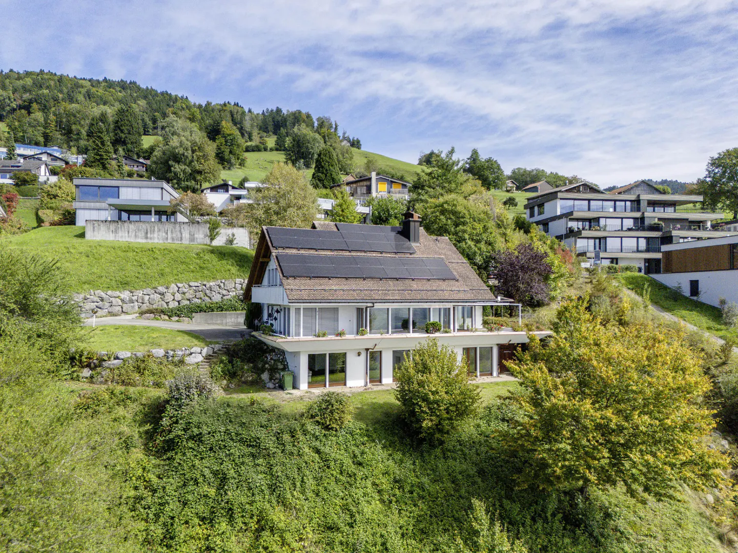 A modern two-story house with solar panels on a green hillside under a blue sky.