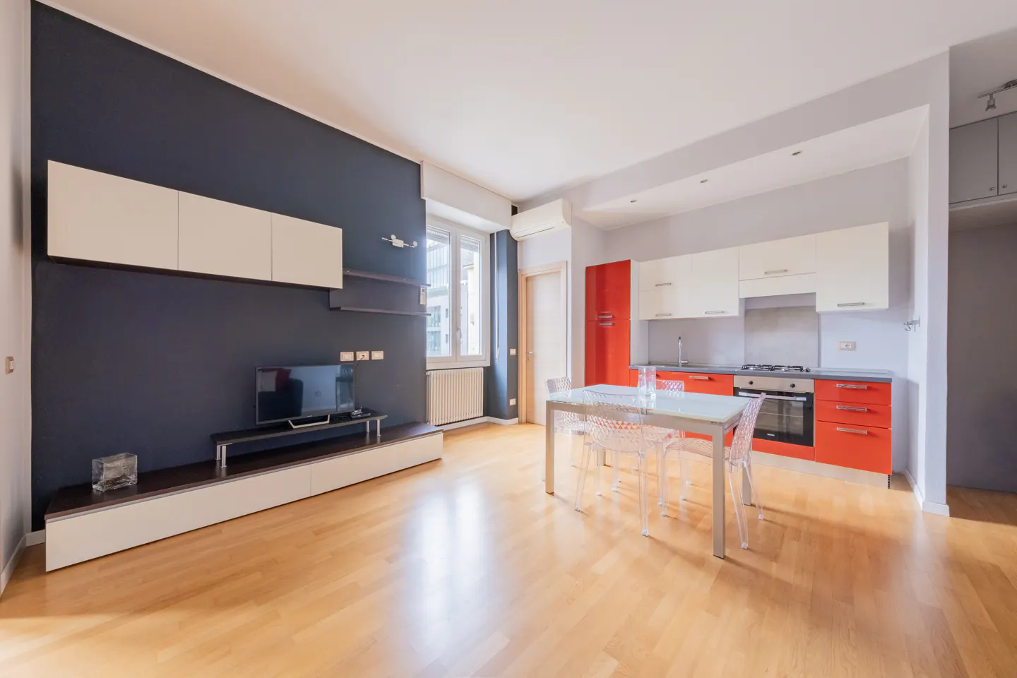 A modern apartment interior with a dark blue accent wall, TV, white cabinets, and a kitchen with orange cabinets. A glass table sits on a light wood floor.