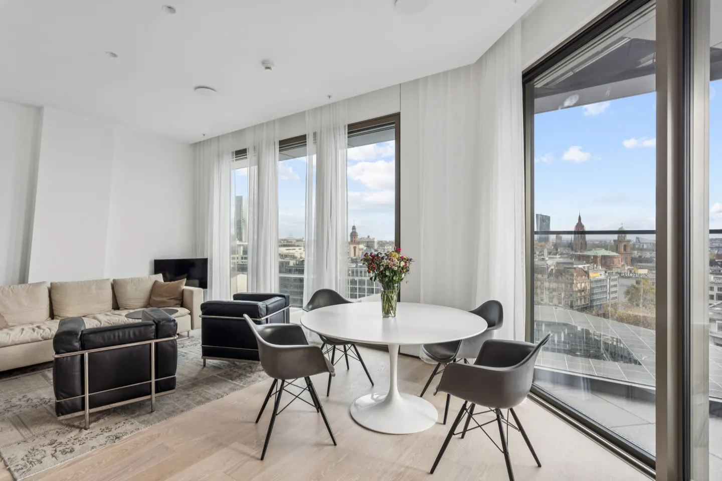 Bright, modern living room with a beige sofa, black chairs, and a round white table with flowers. Large windows offer a city view.
