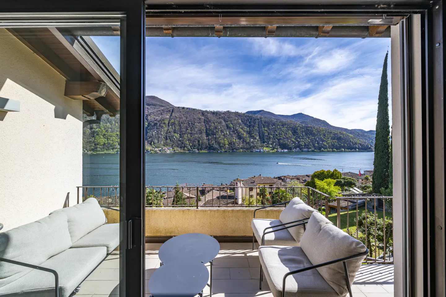 Balcony view of Lake Lugano, Switzerland. Gray outdoor furniture sits on a tan balcony overlooking the lake and mountains.