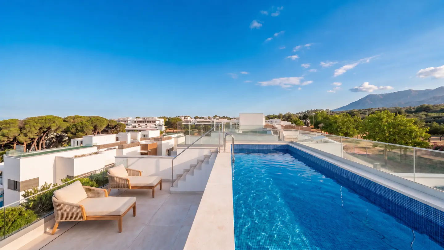 Rooftop view of a blue tiled pool, two lounge chairs, and stairs. White buildings, trees, and mountains are in the background.