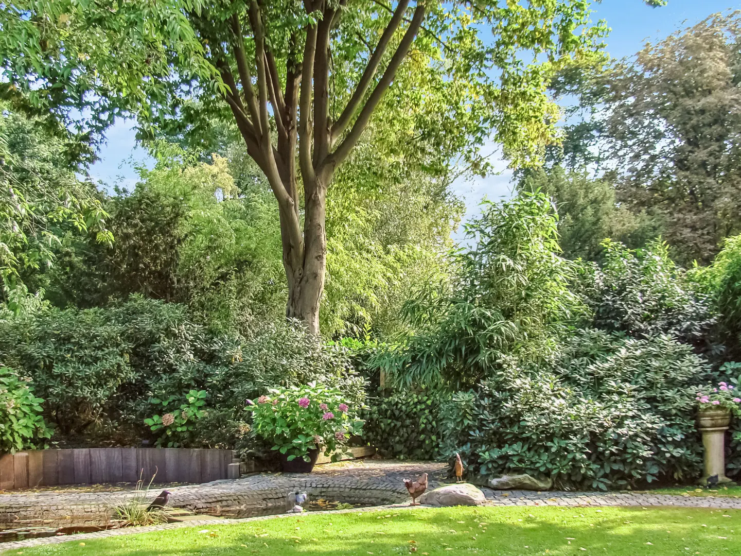 Lush green garden with a small pond, trees, bushes, and flowers under a blue sky. Birds are near the pond.