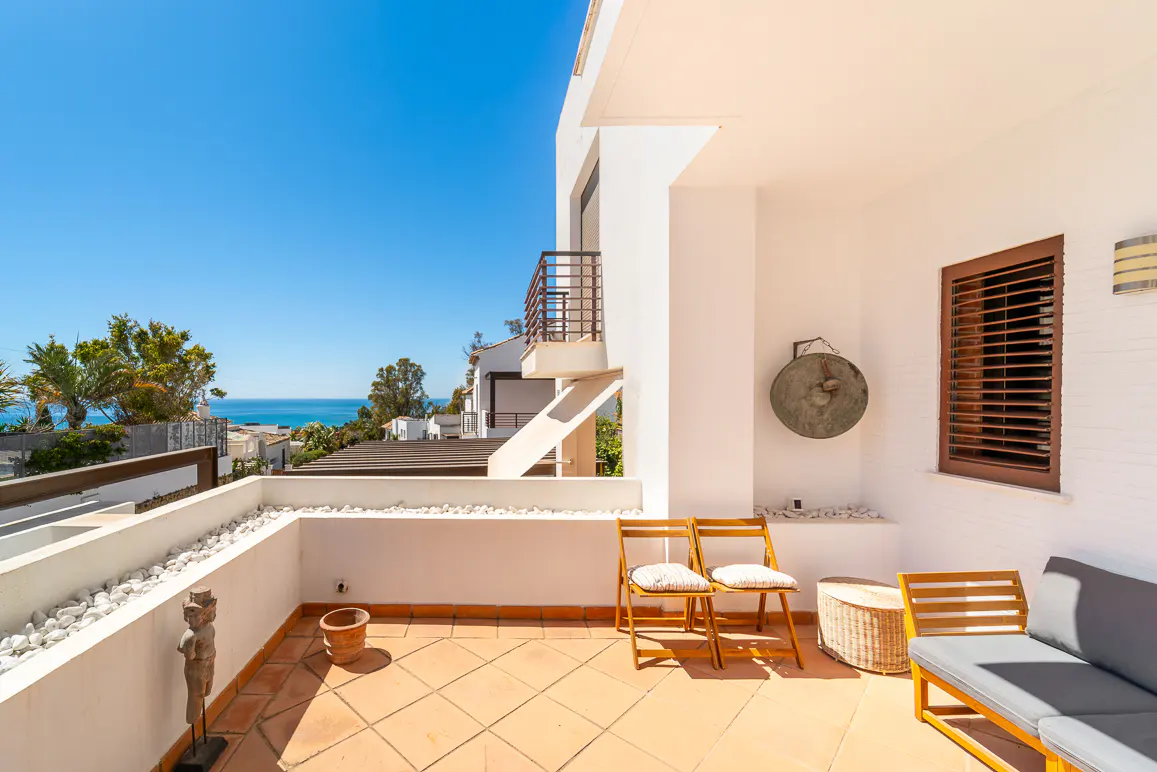 Sunny patio with ocean view. Two wooden chairs, a gray sofa, and a gong decorate the tiled space.
