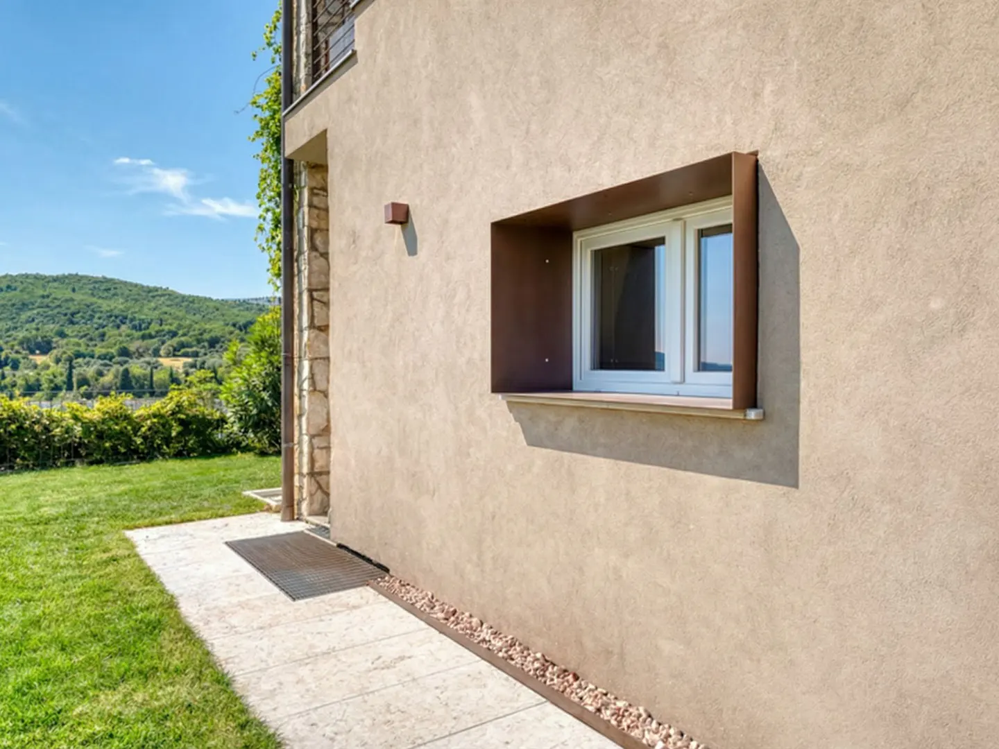 Exterior view of a beige stucco house with a white window framed in brown. A stone walkway leads to a green lawn and a distant, tree-covered hill.