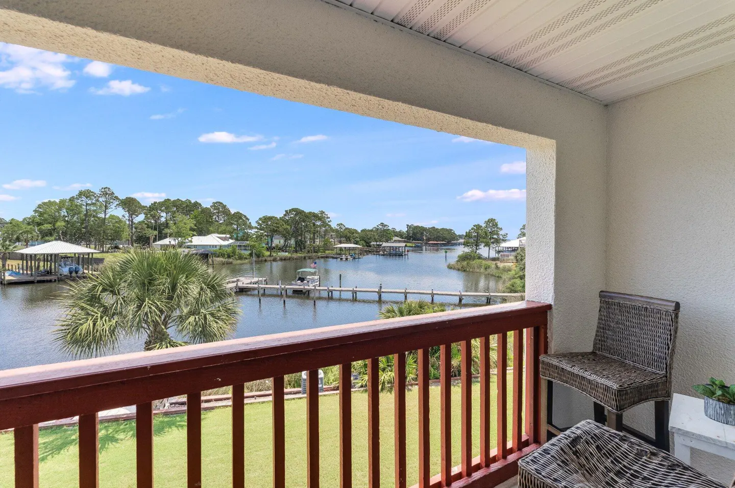 Balcony view of a waterway with docks and boats under a blue sky. Wicker chairs sit on the balcony with a red railing.