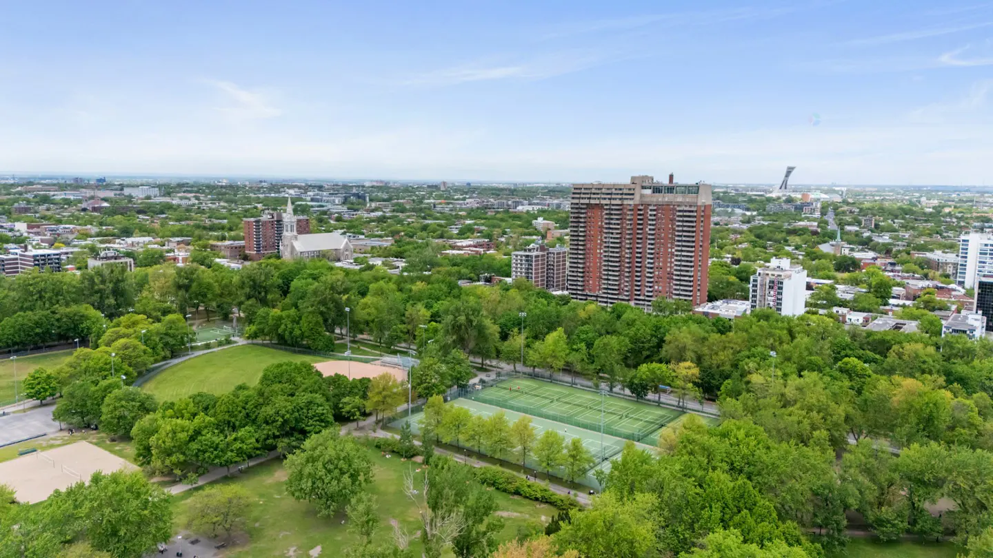 Aerial view of a lush green park with tennis courts, surrounded by city buildings, a church, and a clear blue sky.