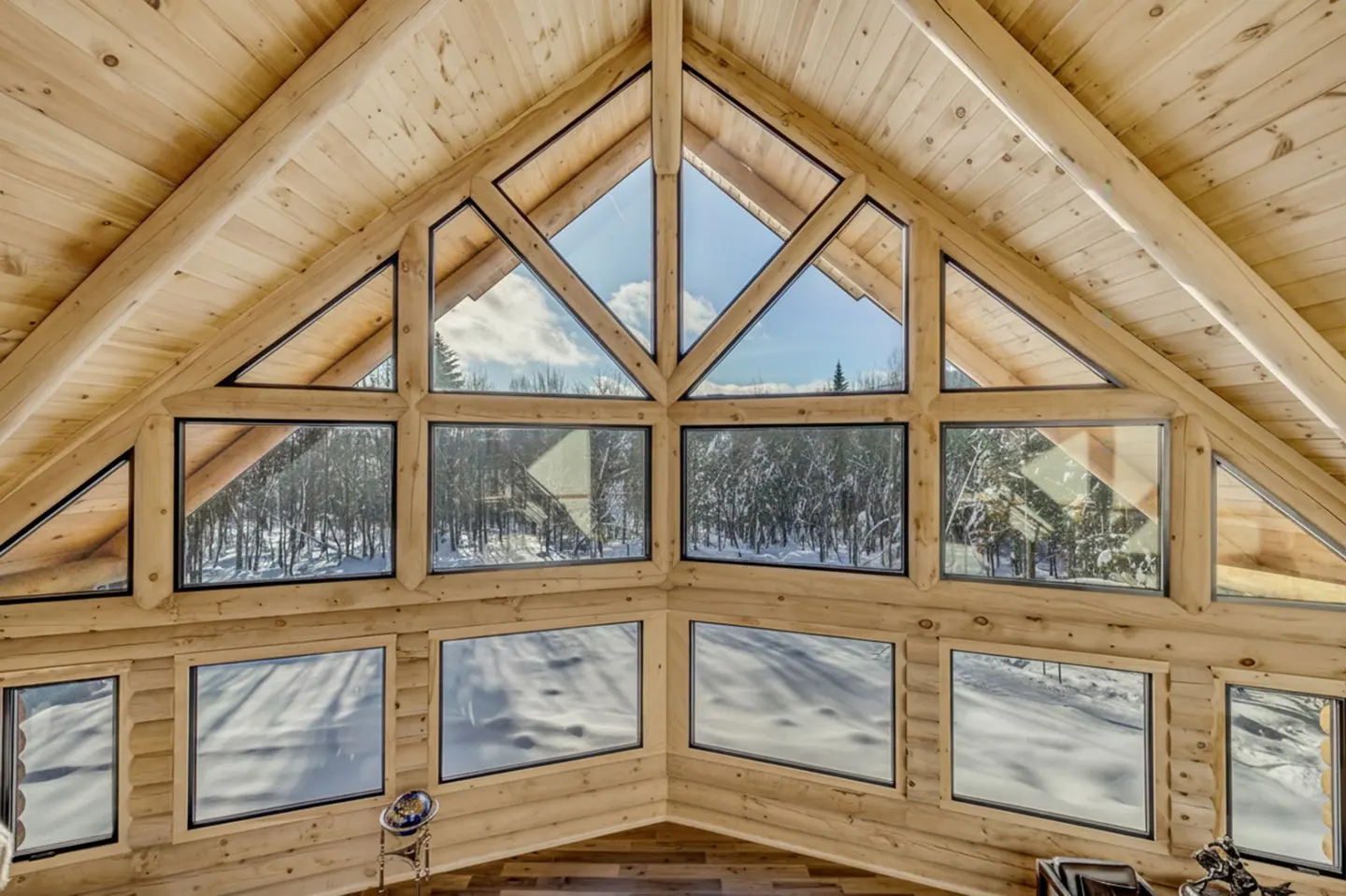 Interior view of a log cabin with a wall of windows showing a snowy forest. The windows are framed in black and the walls are light wood.