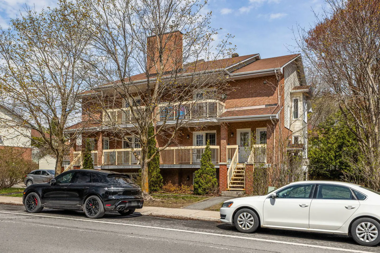 Two-story brick apartment building with balconies, brown roof, and chimney. A black SUV and white sedan are parked on the street.
