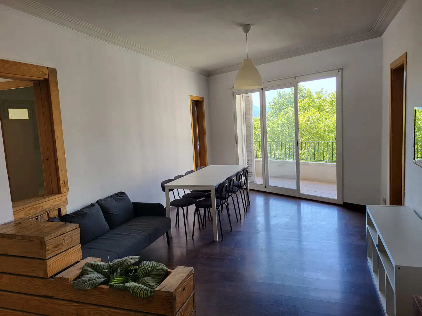 Bright living room with white walls, dark wood floors, a long white table with black chairs, and a gray sofa. Balcony doors open to green trees.