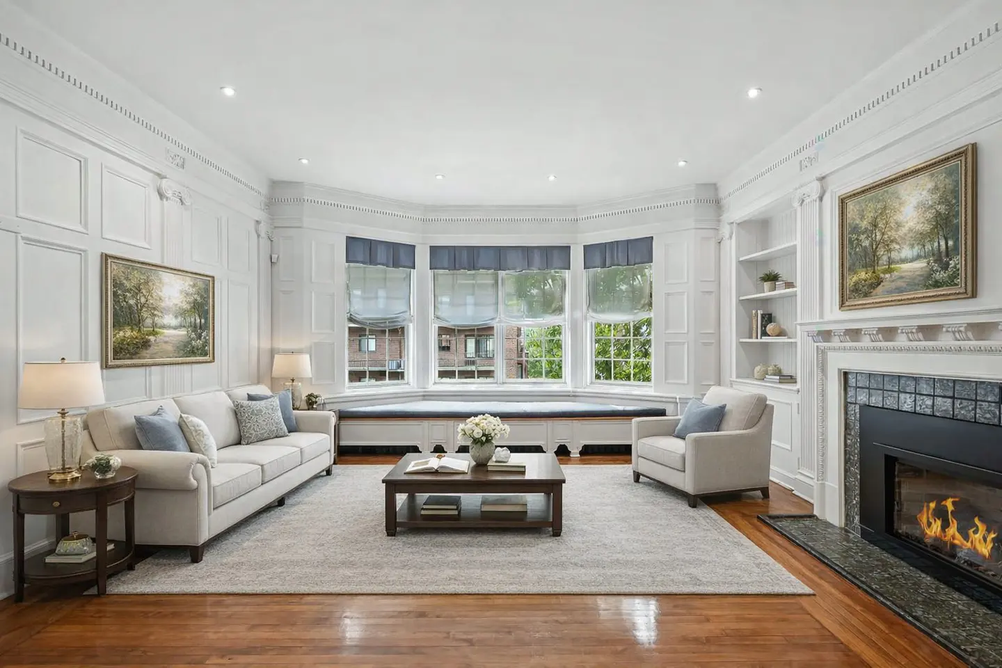 Bright living room with white walls, hardwood floors, and a fireplace. A sofa, chair, and bay window with blue shades are visible.